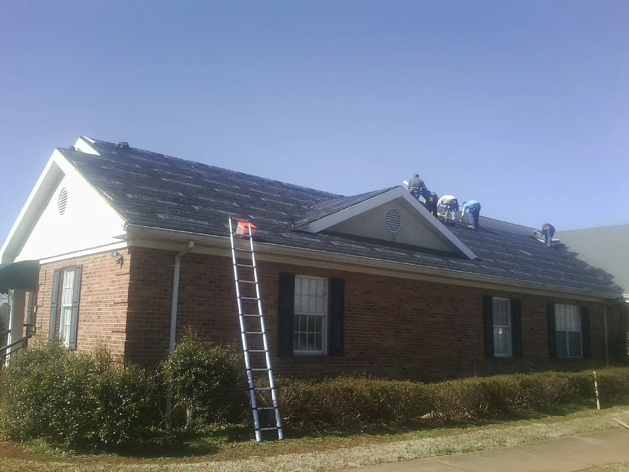 Crew tearing shingles off a brick ranch roof with a ladder set — Emergency Roof Repair work by our crew in Conklin