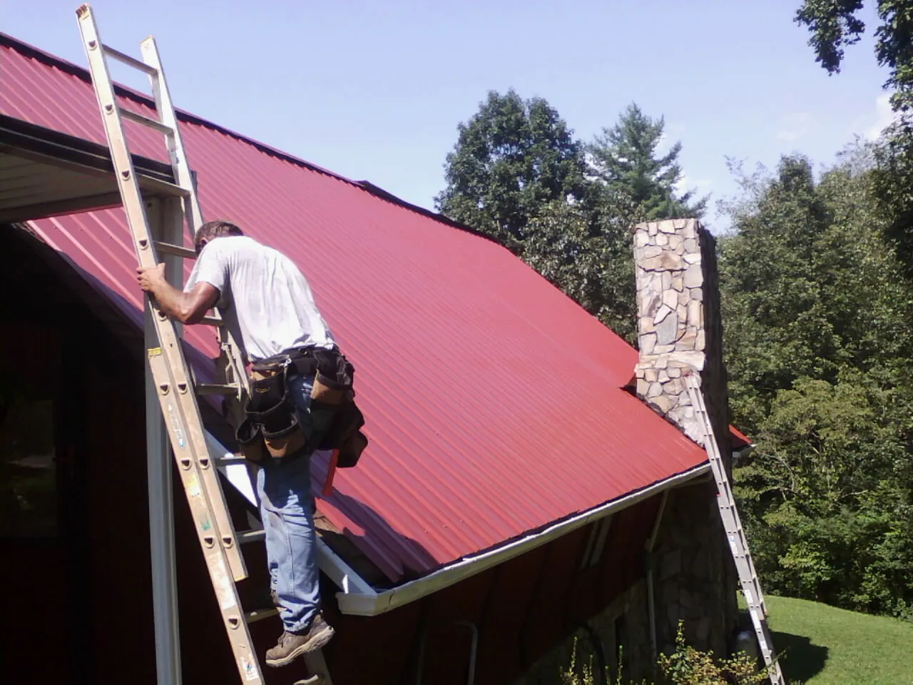 Roofer on a ladder beside a red metal roof and stone chimney — Soffit & Fascia Repair by Roofing Brothers in Conklin